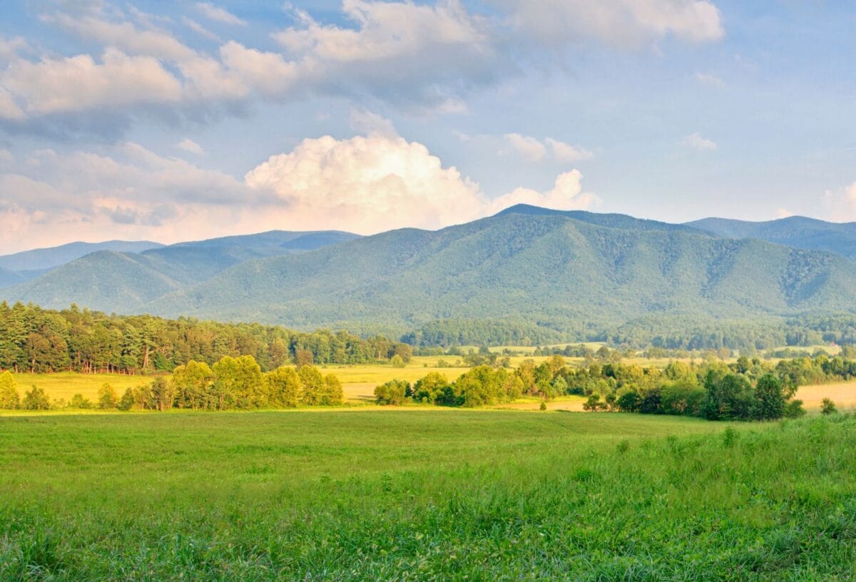 A wide scenic view of the lush green valley at Cades Cove in Great Smoky Mountains National Park, featuring rolling mountains under a cloudy blue sky at sunset