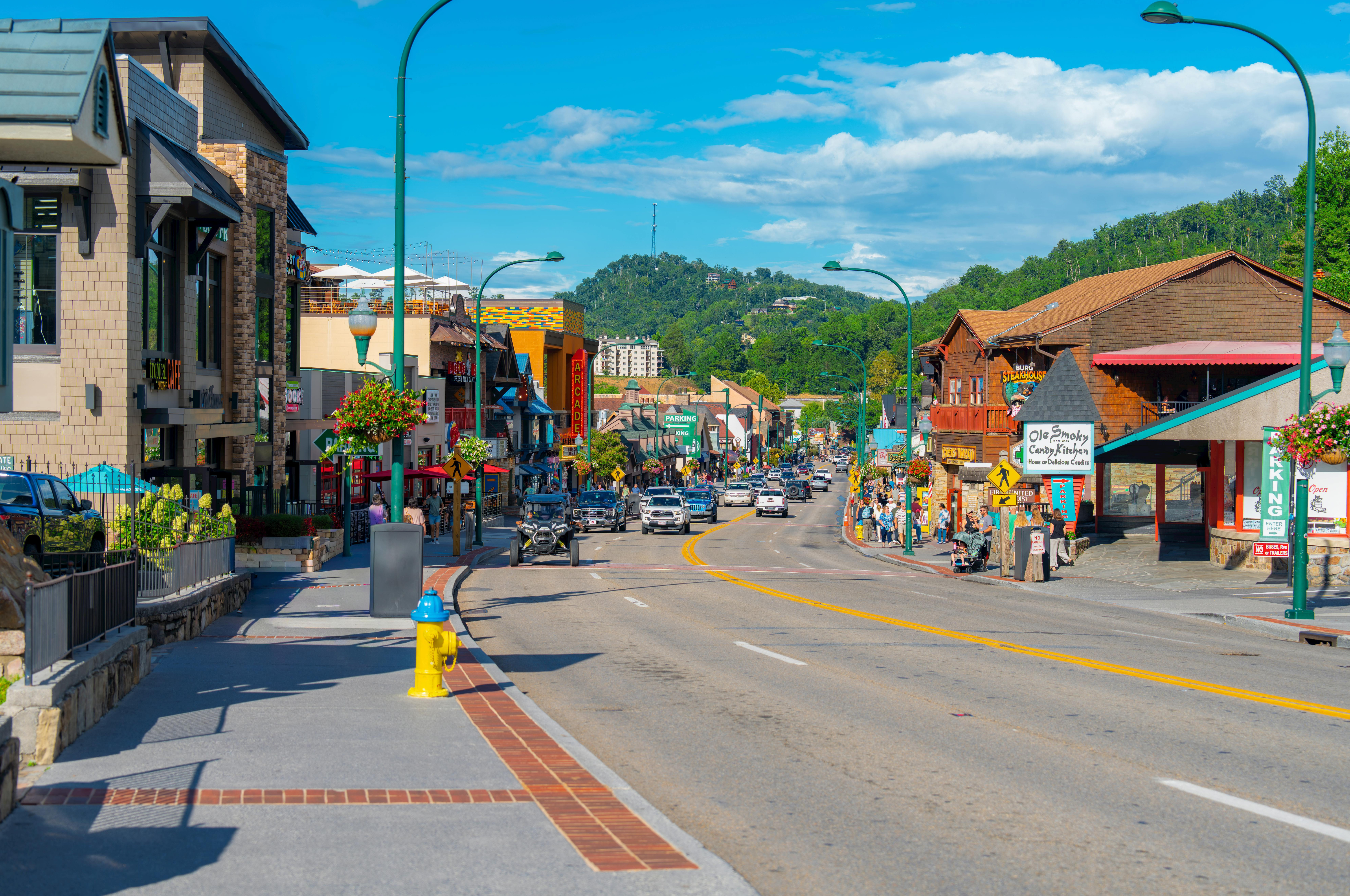 Main street in Gatlinburg, Tennessee, on a sunny day with cars and pedestrians, looking towards the tree-covered mountains in the background. The street is lined with shops, including the 'Old Smoky Candy Kitchen', and features a blue and yellow fire hydrant in the foreground.