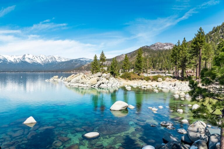 Scenic view of granite boulders and crystal clear turquoise water at North Shore Lake Tahoe with snow-capped mountains in the background.