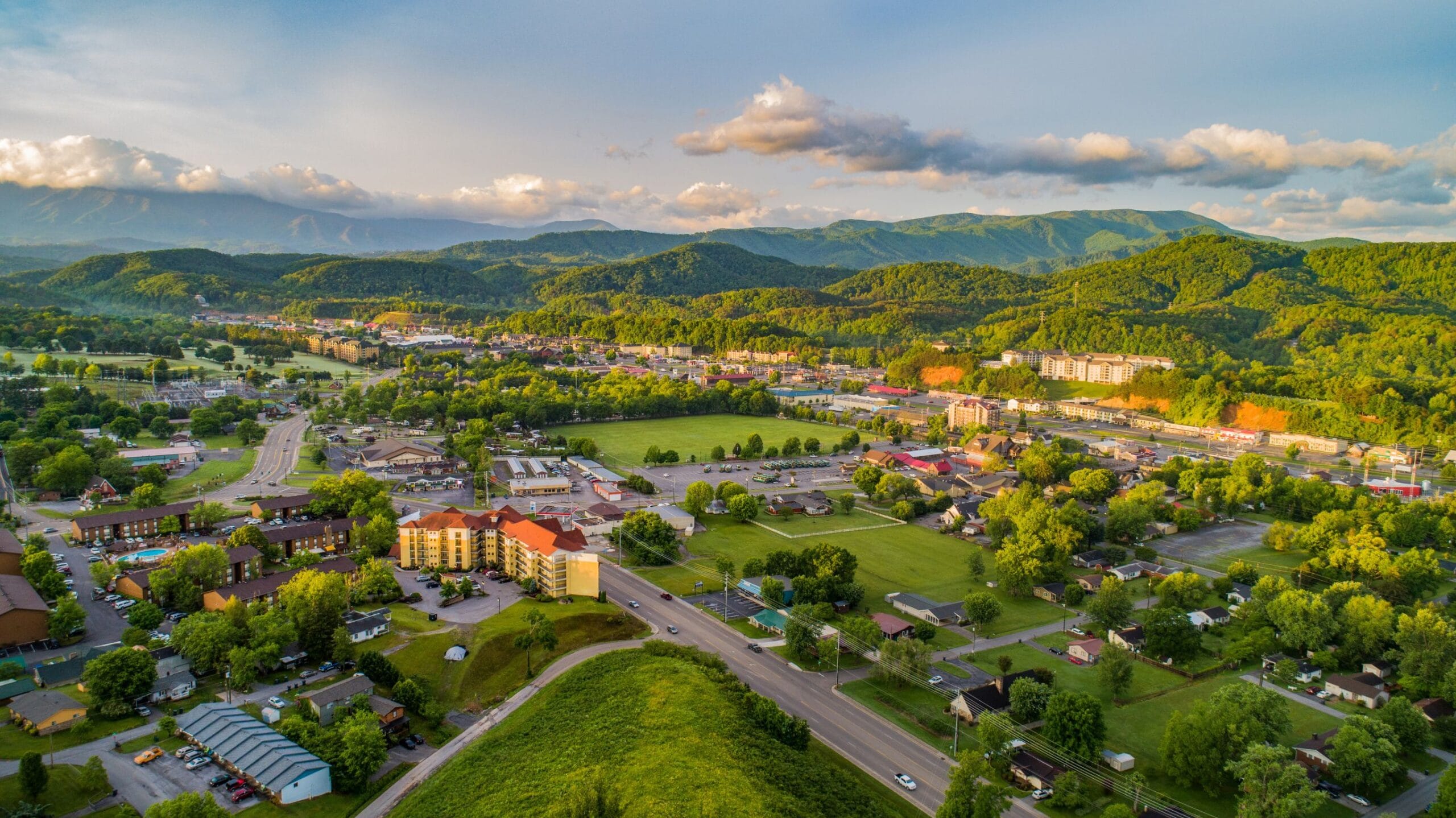 Aerial view of Pigeon Forge and Sevierville, Tennessee, showing hotels, roads, and green spaces nestled against the rolling green foothills of the Great Smoky Mountains.