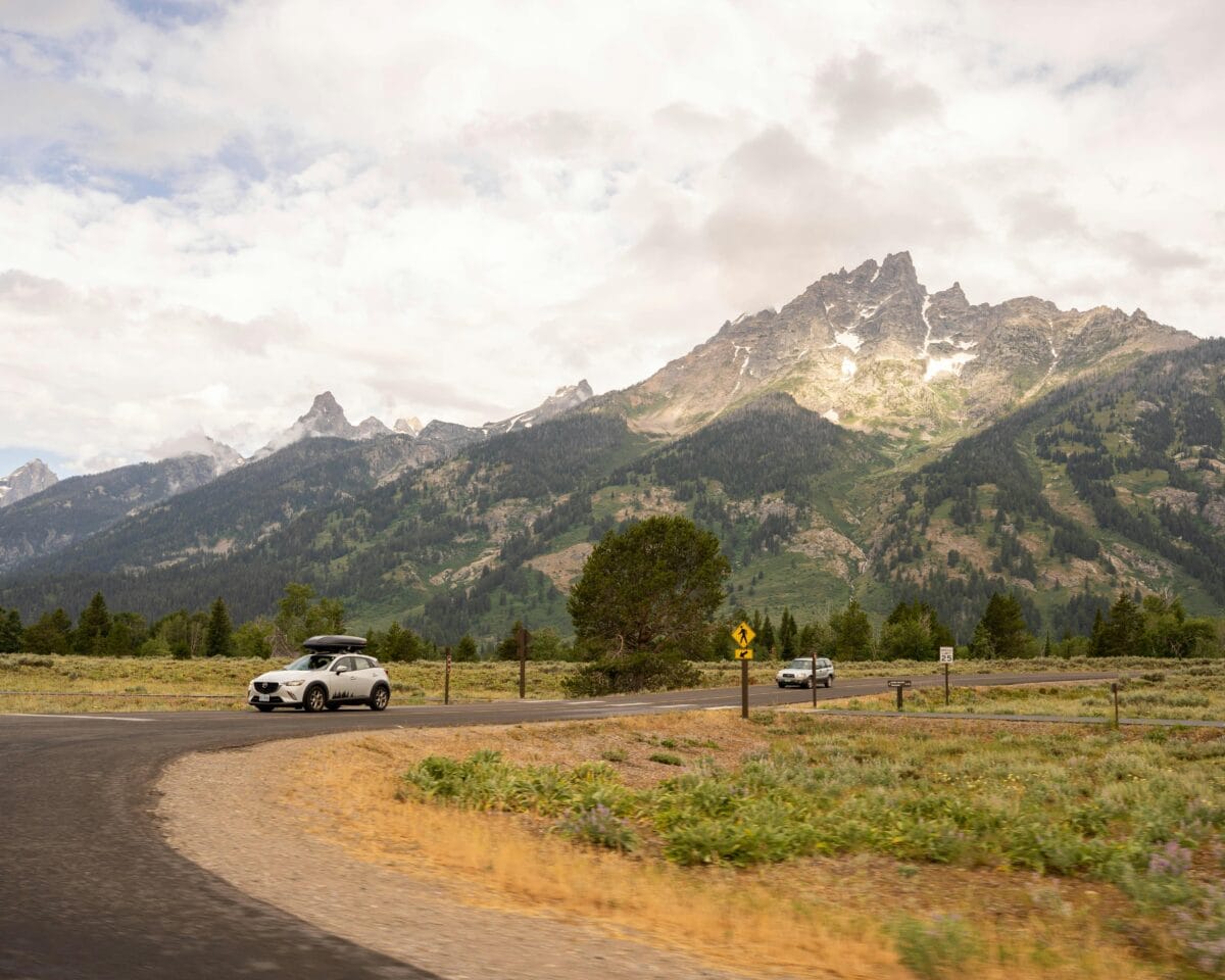 A SUV with cargo box on top on a roas with a scenic moutain in the backgorund.