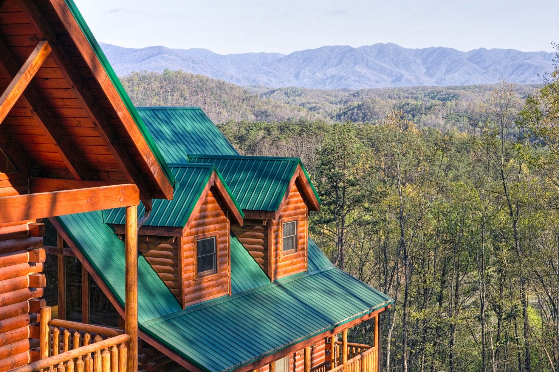 Close-up view of a multi-level log cabin with a green metal roof and wooden balconies, overlooking a lush forest and the rolling blue ridges of the Great Smoky Mountains.