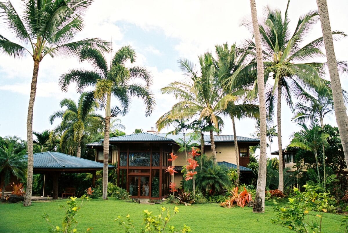 Large luxury vacation rental home on the Big Island's Kohala Coast, featuring high ceilings, dark wood trim, and a manicured green lawn surrounded by tropical palm trees.