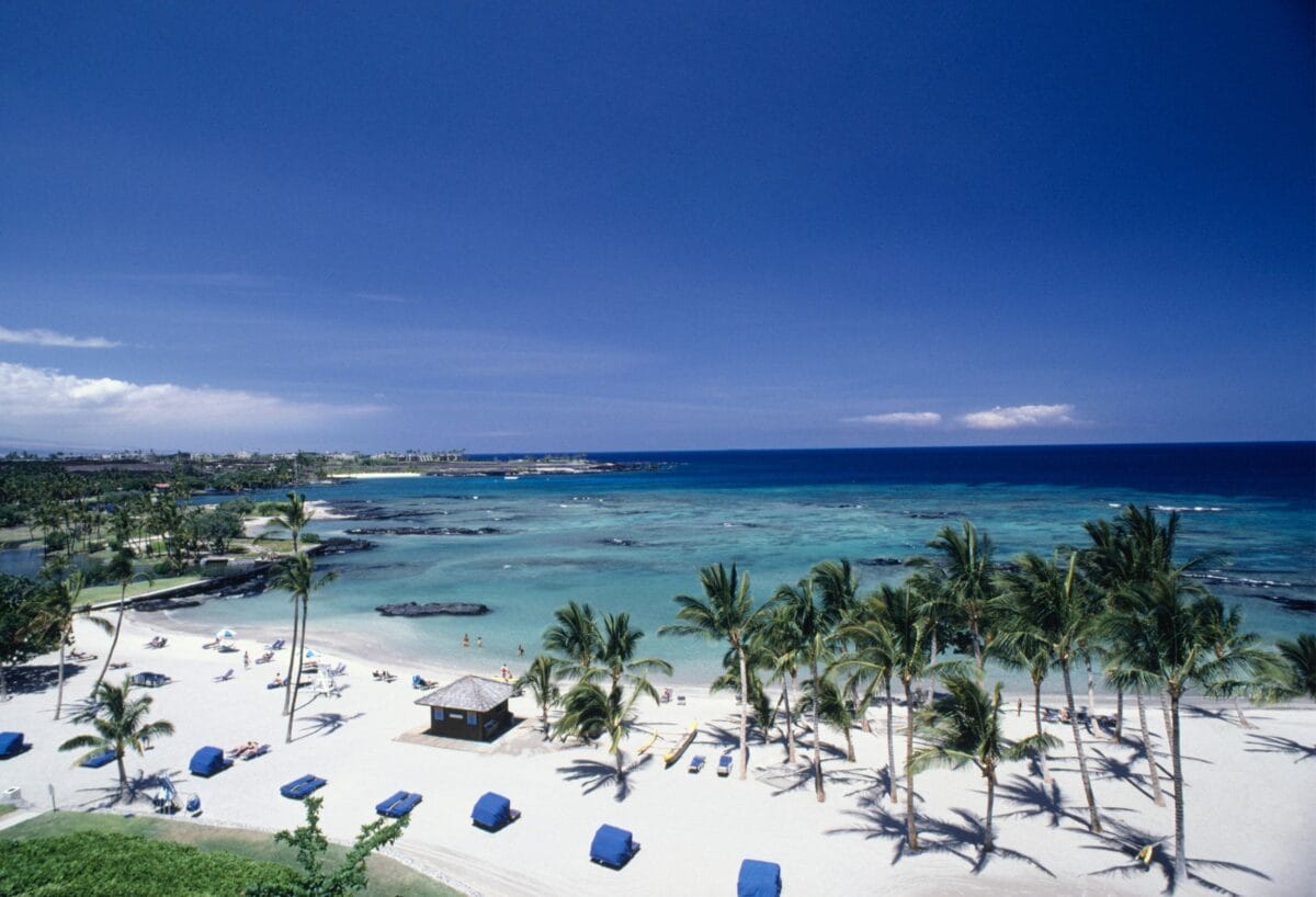 High-angle view of the white sand beach and calm turquoise water at Mauna Lani Bay on the Kohala Coast, showing palm trees, beach umbrellas, and the protected reef area.