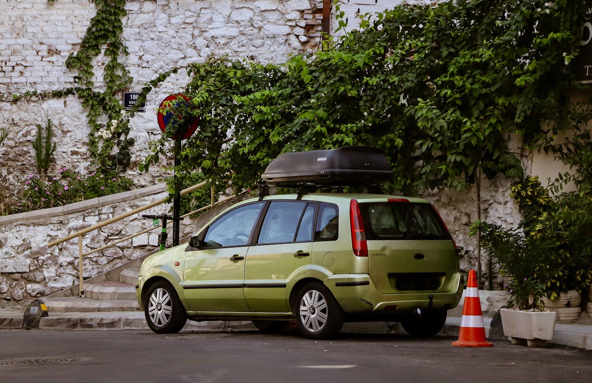 A green car with rooftop box parked on a leafy street in İzmir, Türkiye, against a brick wall.