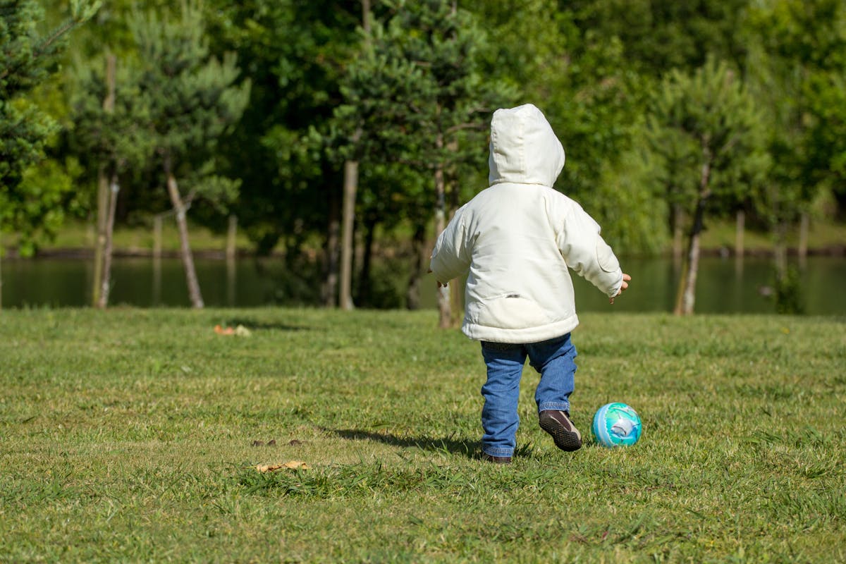 A small child in a coat plays football on a sunny day in a lush green park.