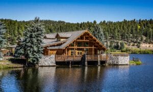 A traditional dark wood log cabin situated on a rocky shoreline with clear alpine water and pine trees, evoking a nostalgic family cottage atmosphere at Lake Tahoe.