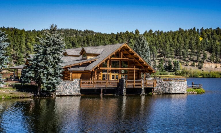 A traditional dark wood log cabin situated on a rocky shoreline with clear alpine water and pine trees, evoking a nostalgic family cottage atmosphere at Lake Tahoe.